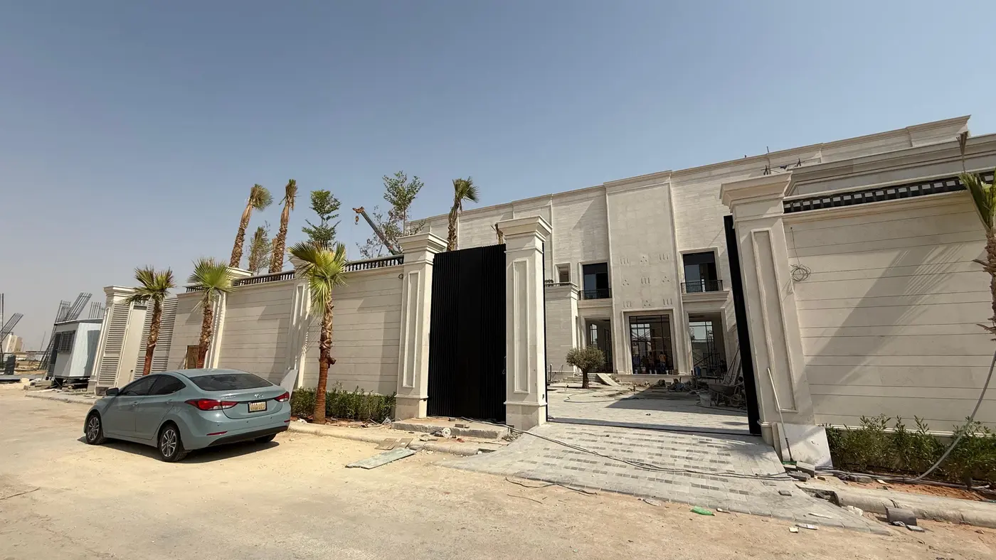 The wall and gated entrance of a stone-clad compound fronted by palm trees